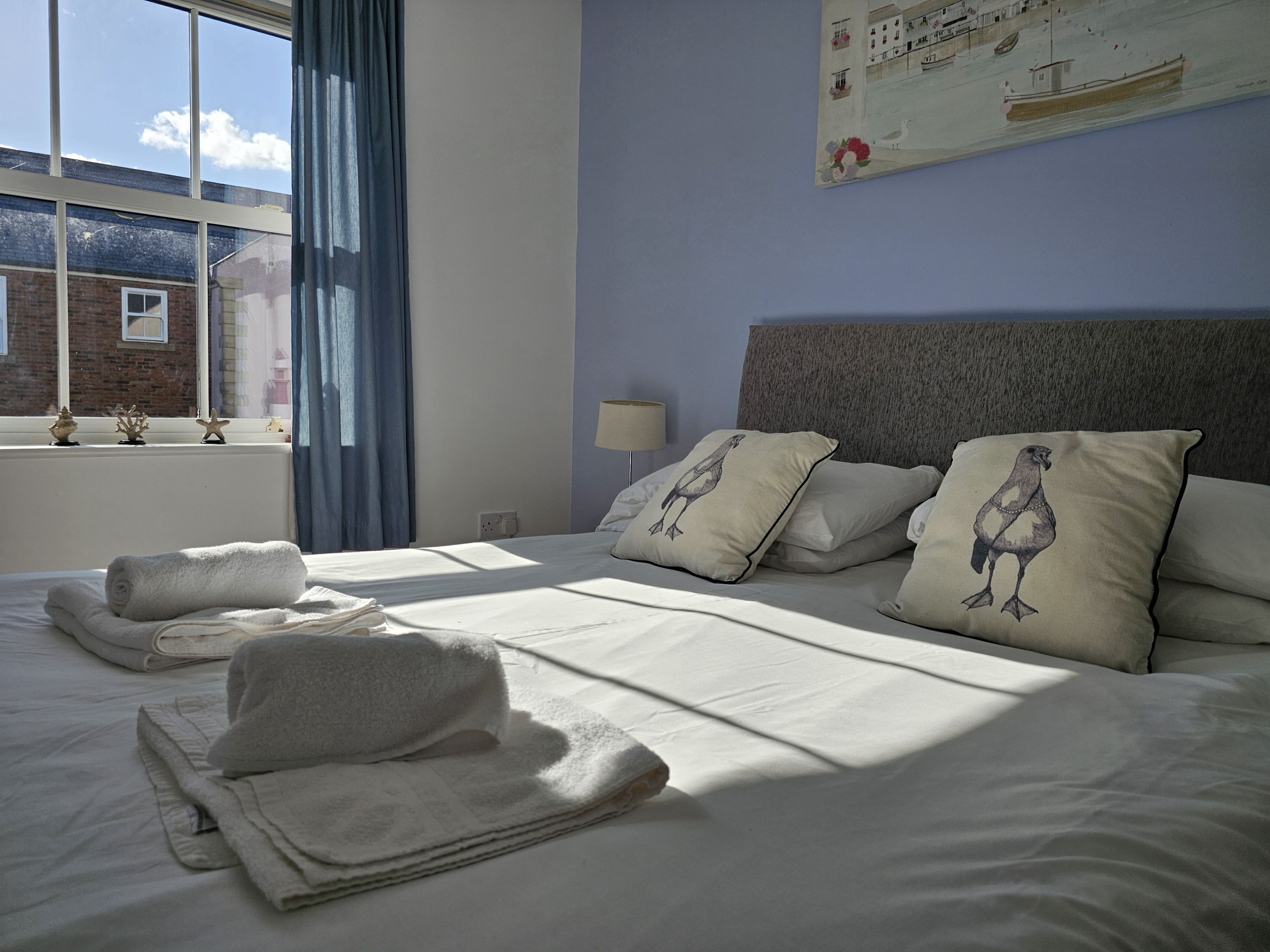 Et hotelværelse med en pænt redt seng, et vindue med gardiner, en lampe og et billede på væggen.