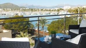 Un balcon agrémenté de meubles en osier, une table avec des tasses et un menu, et un port de plaisance avec des bateaux en arrière-plan.