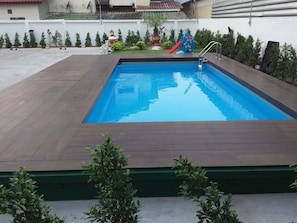 Una piscina rettangolare con bordo in legno, circondata da vegetazione e uno scivolo.
