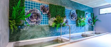 Un área de piscina interior moderna con una pared verde, flores decorativas y una fuente de agua cristalina.