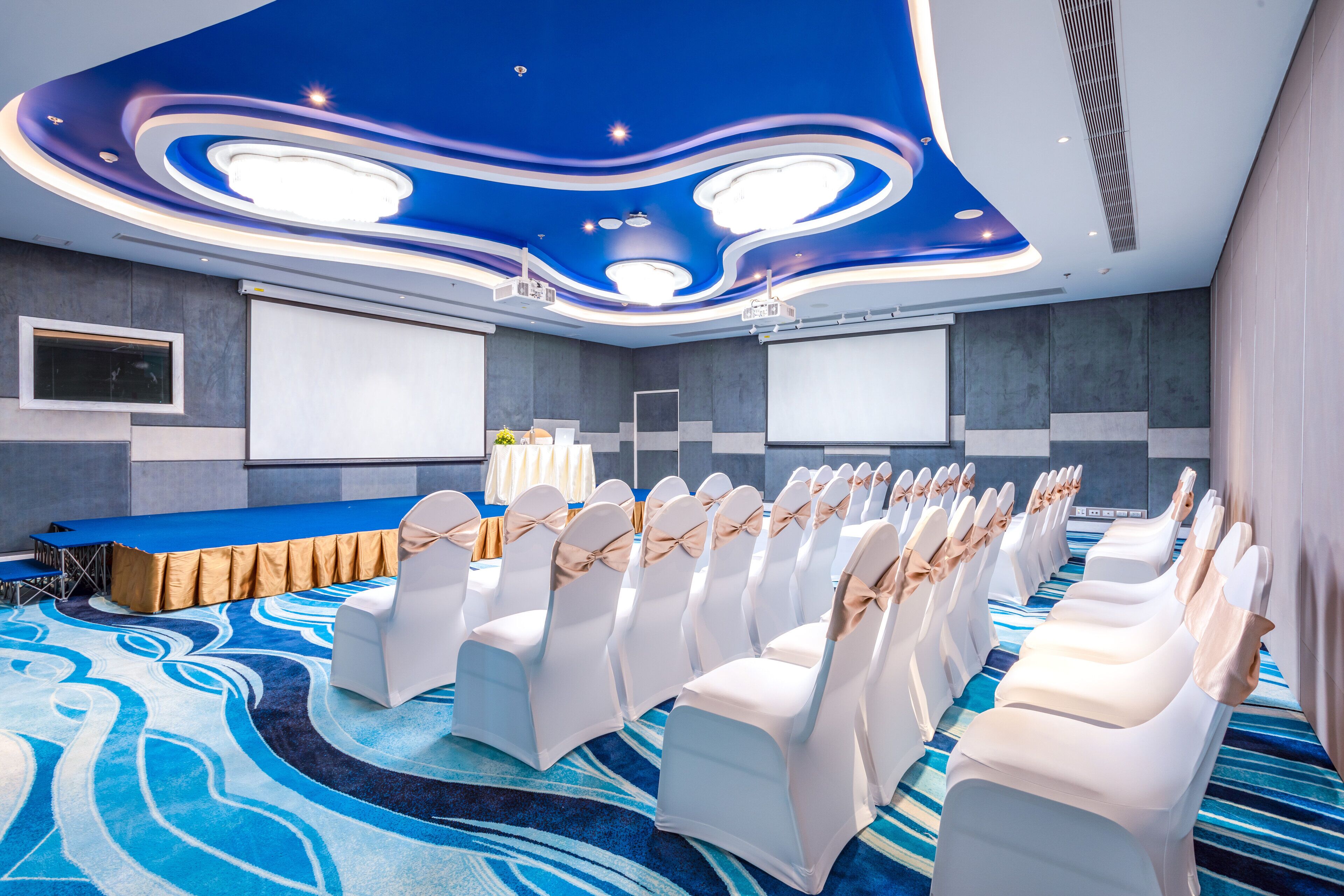 Een congreszaal met een podium, rijen witte stoelen versierd met linten, een blauw tapijt en een modern plafondontwerp.