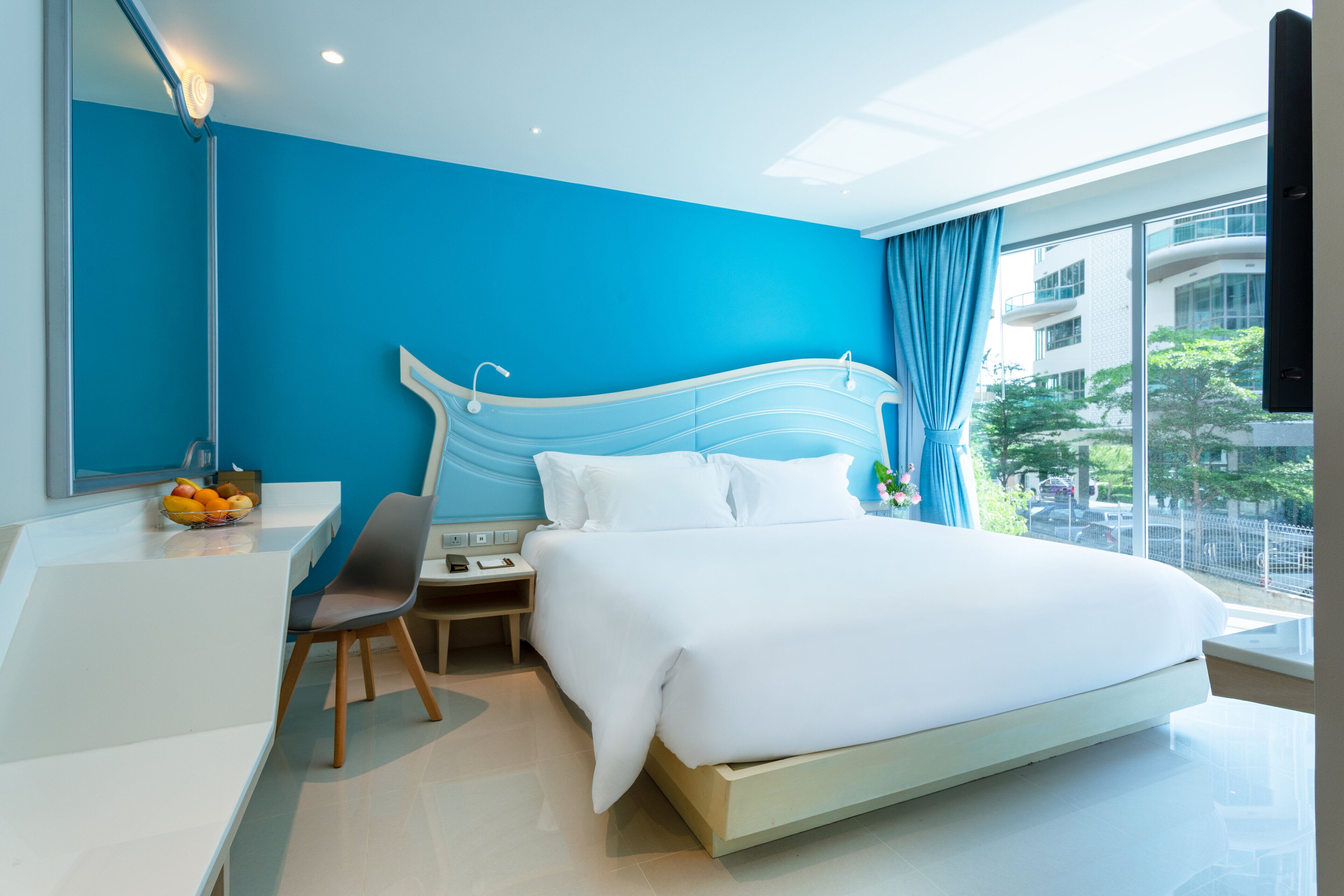 A modern hotel room with a large bed, a desk with a chair, and a view of the city through large windows.