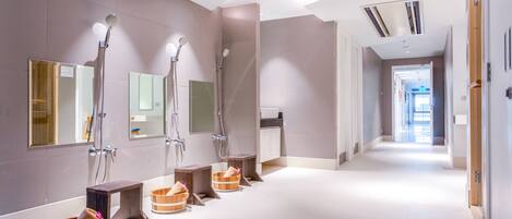 A modern bathroom with multiple showerheads, wooden bathtubs, and a long hallway leading to a reception area.