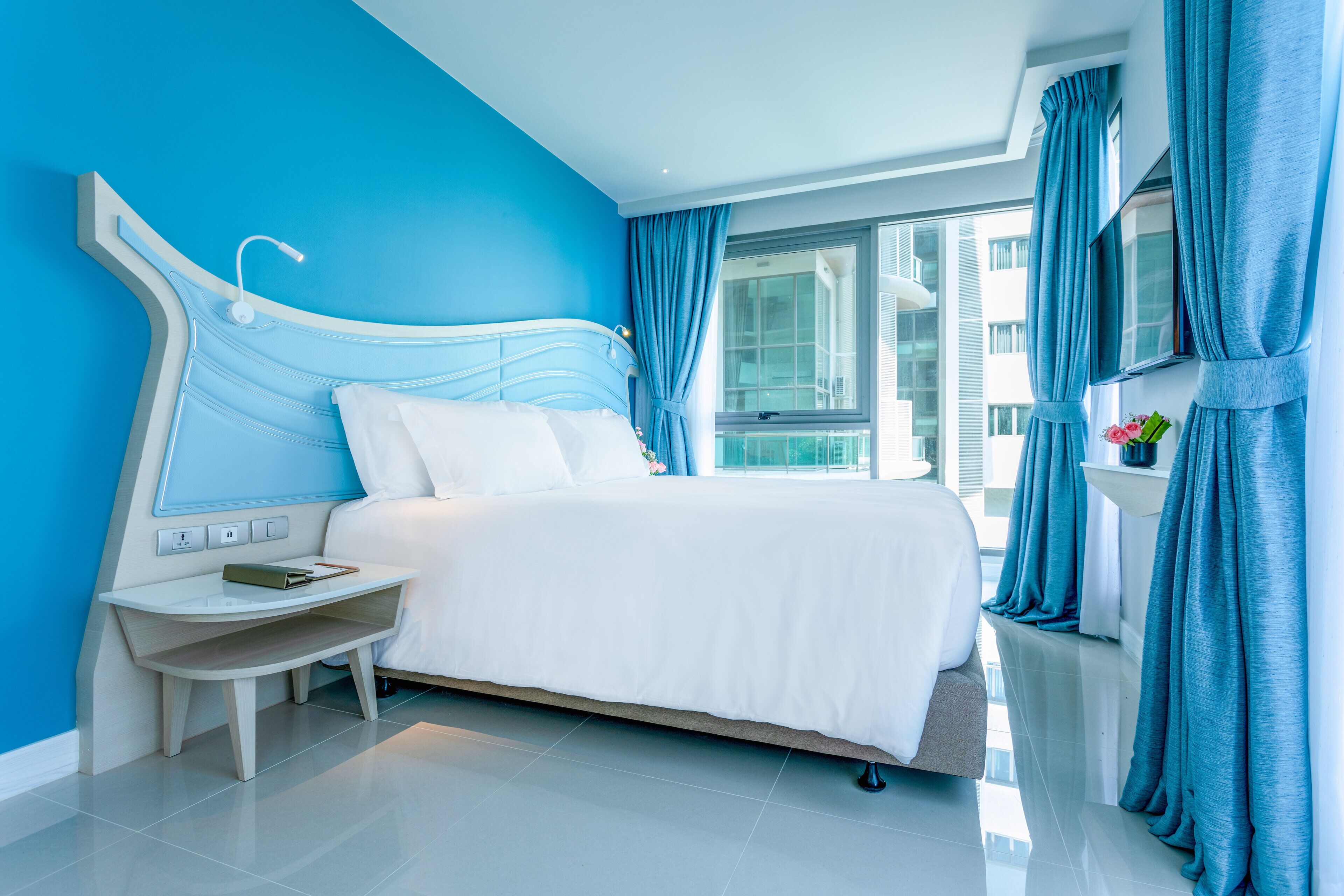 A modern hotel room with a large bed, a bedside table with a lamp, and a view of the city through large windows.