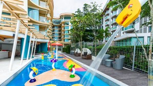 Una piscina con patrones de olas coloridos y fuentes de agua, rodeada de edificios modernos y mobiliario exterior.