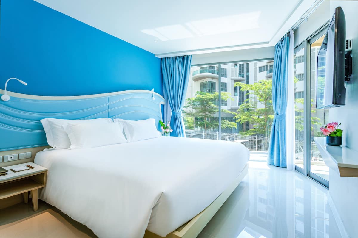 Ett modernt hotellrum med en stor säng, ett nattduksbord och utsikt över staden genom stora fönster.