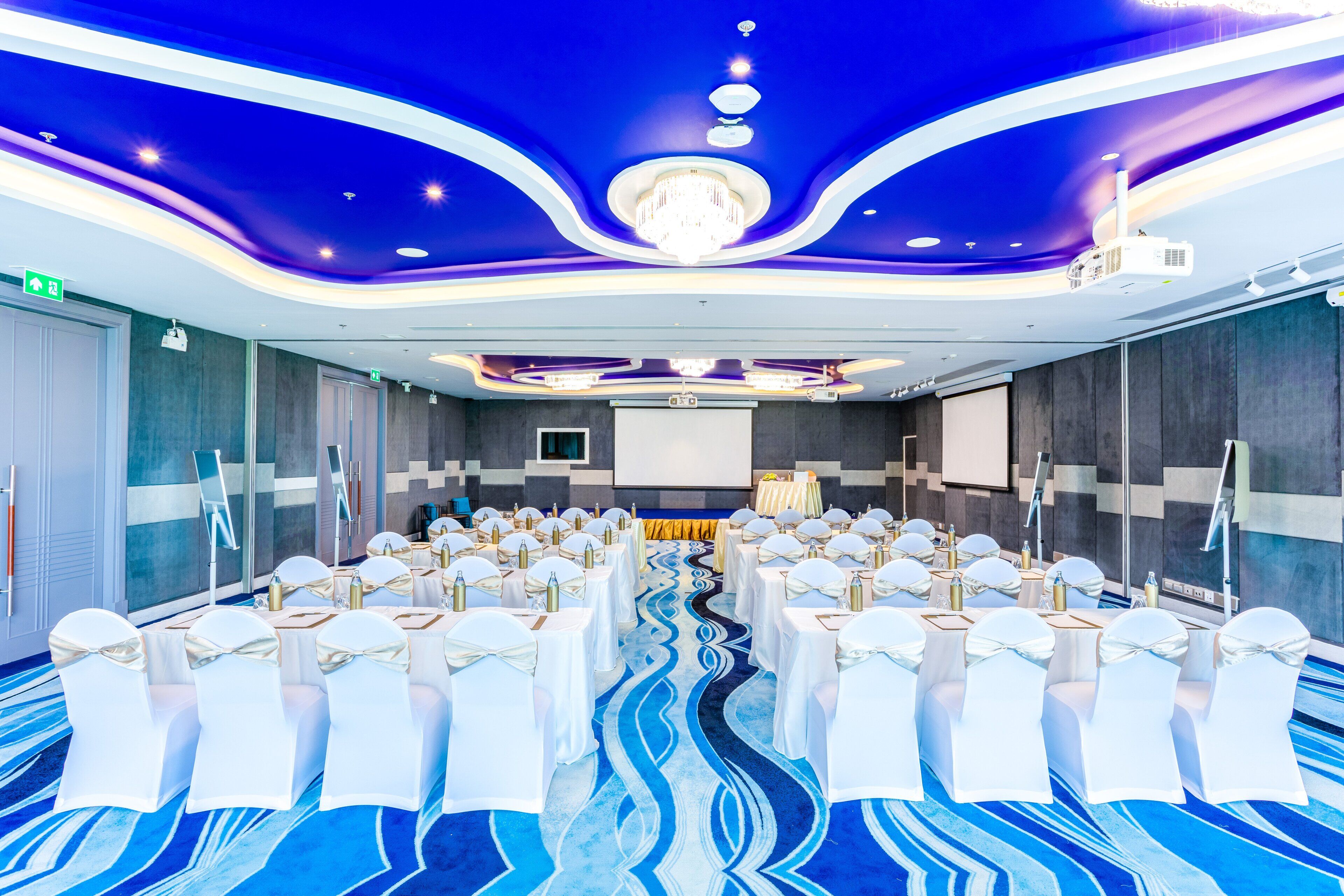 Un salón de conferencias con filas de sillas blancas, una alfombra azul y una pantalla grande al frente.