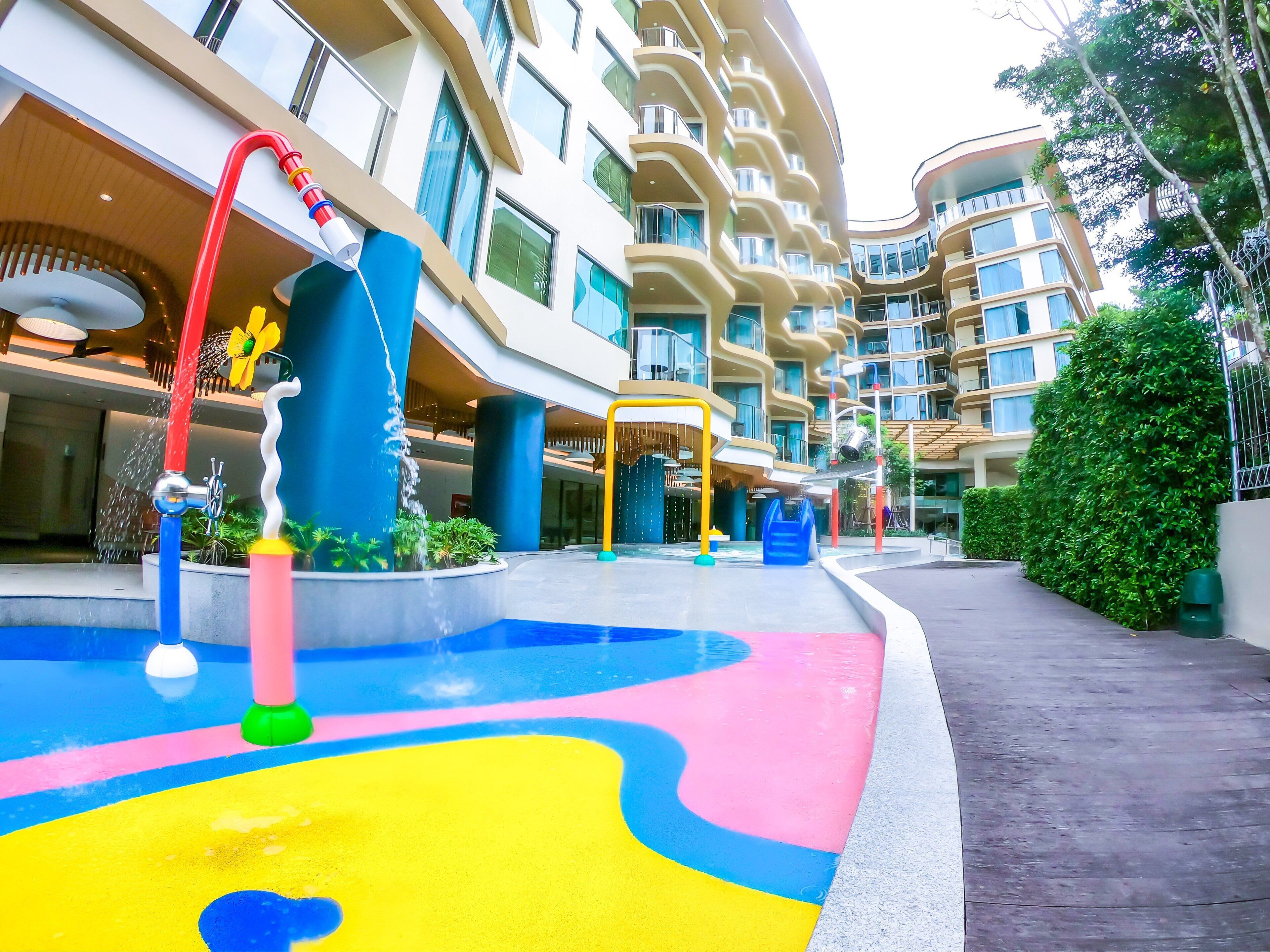 Un área de juegos acuáticos con toboganes de colores y una fuente, rodeada de edificios modernos con balcones y grandes ventanales.