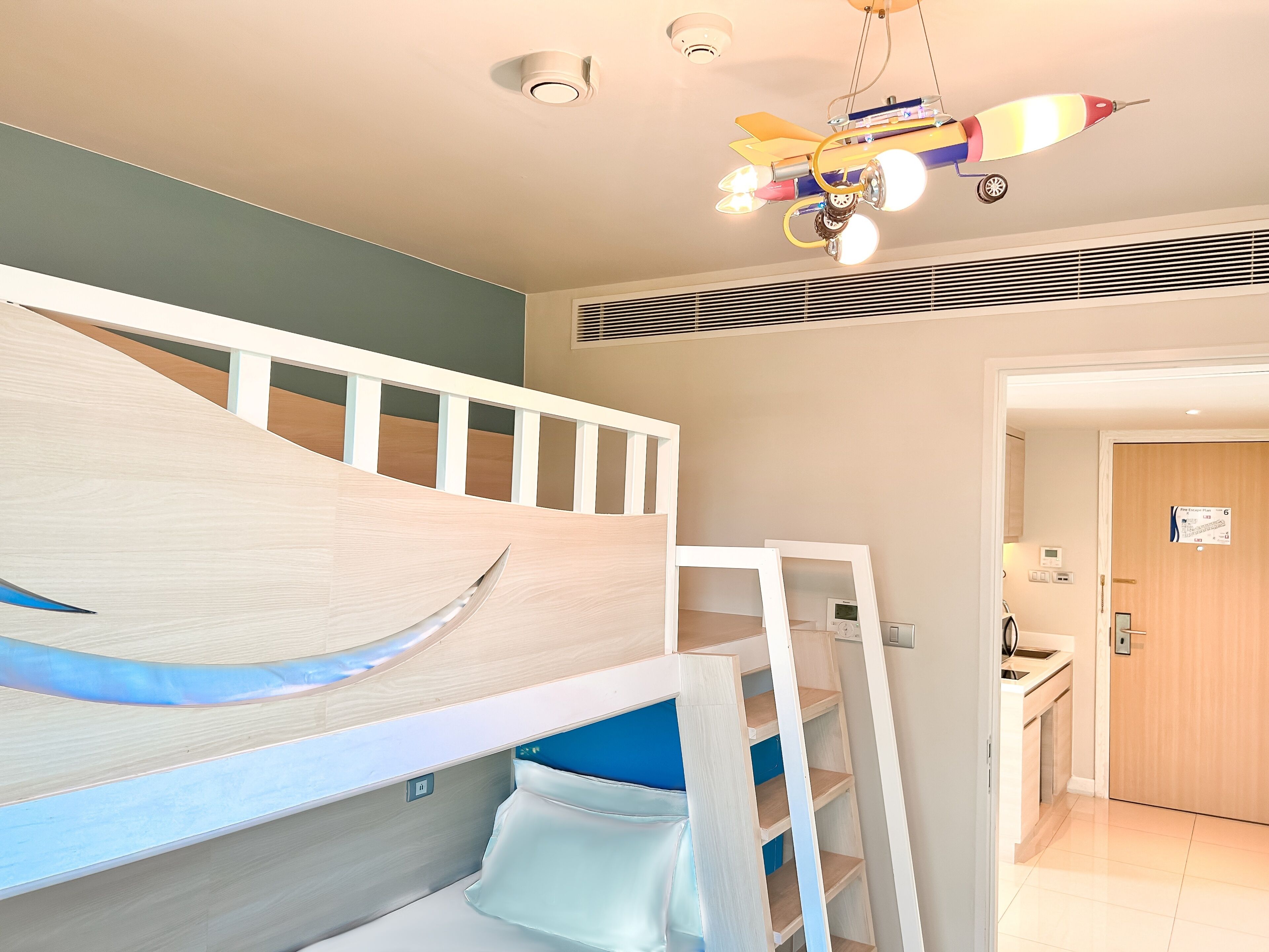 A room with bunk beds, a staircase, a kitchen area, and a ceiling-mounted light fixture.