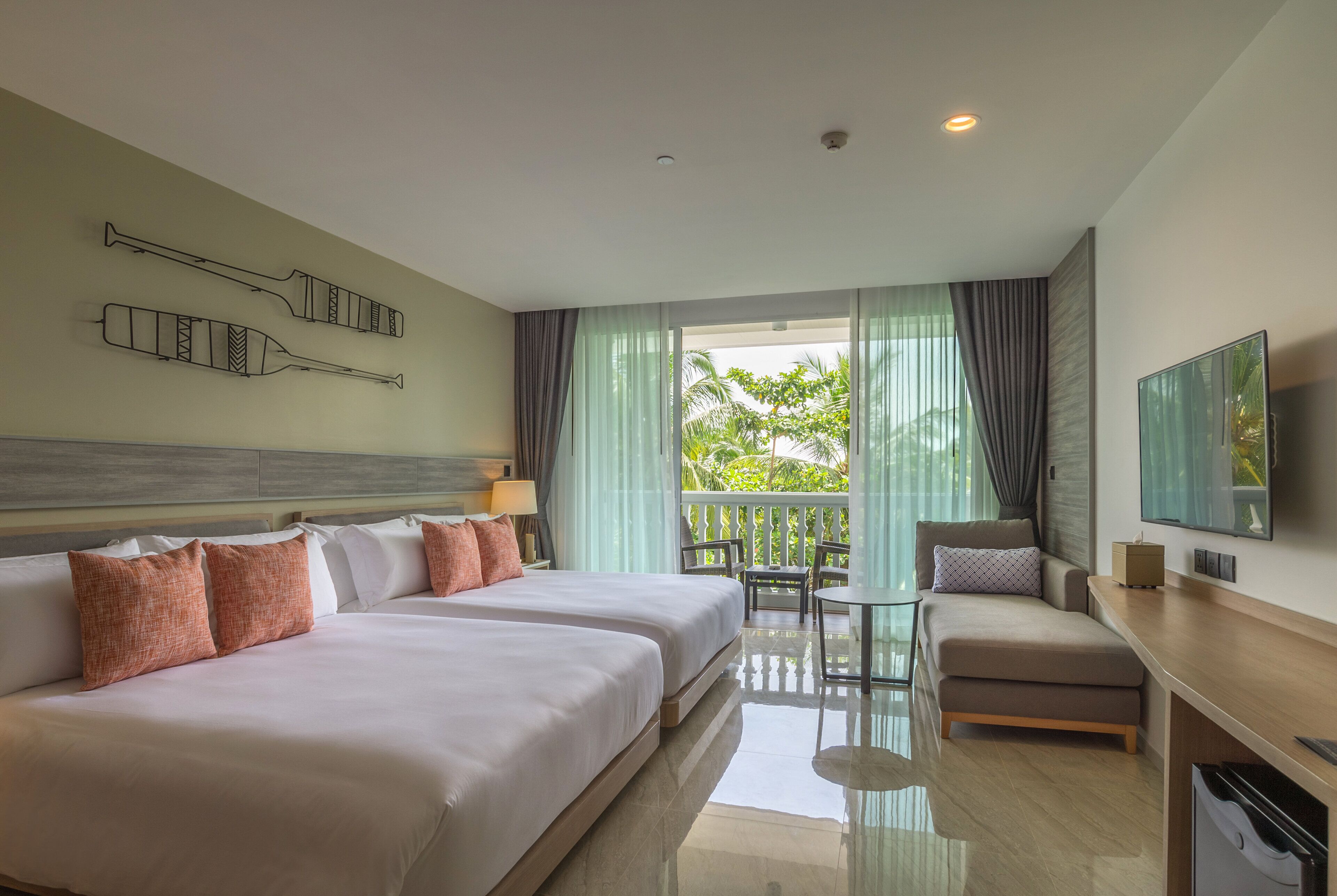 A hotel room with a large bed, a desk, a chair, and a balcony with a view of greenery.