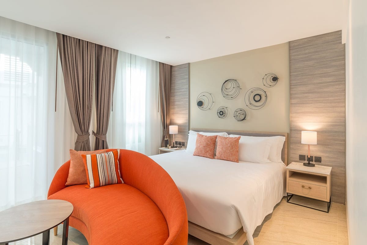 Ein modernes Hotelzimmer mit einem großen Bett, einem gewölbten orangefarbenen Sessel, einem kleinen Tisch und einer Wandlampe.