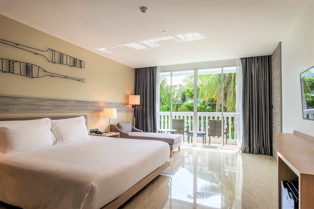 Ein Hotelzimmer mit einem großen Bett, zwei Nachttischen, einem Schreibtisch, einem Sessel und einem Balkon mit Blick auf viel Grün, inklusive einer Deckenleuchte.