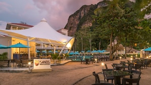 Un club de playa con mobiliario exterior, una piscina y montañas al fondo.