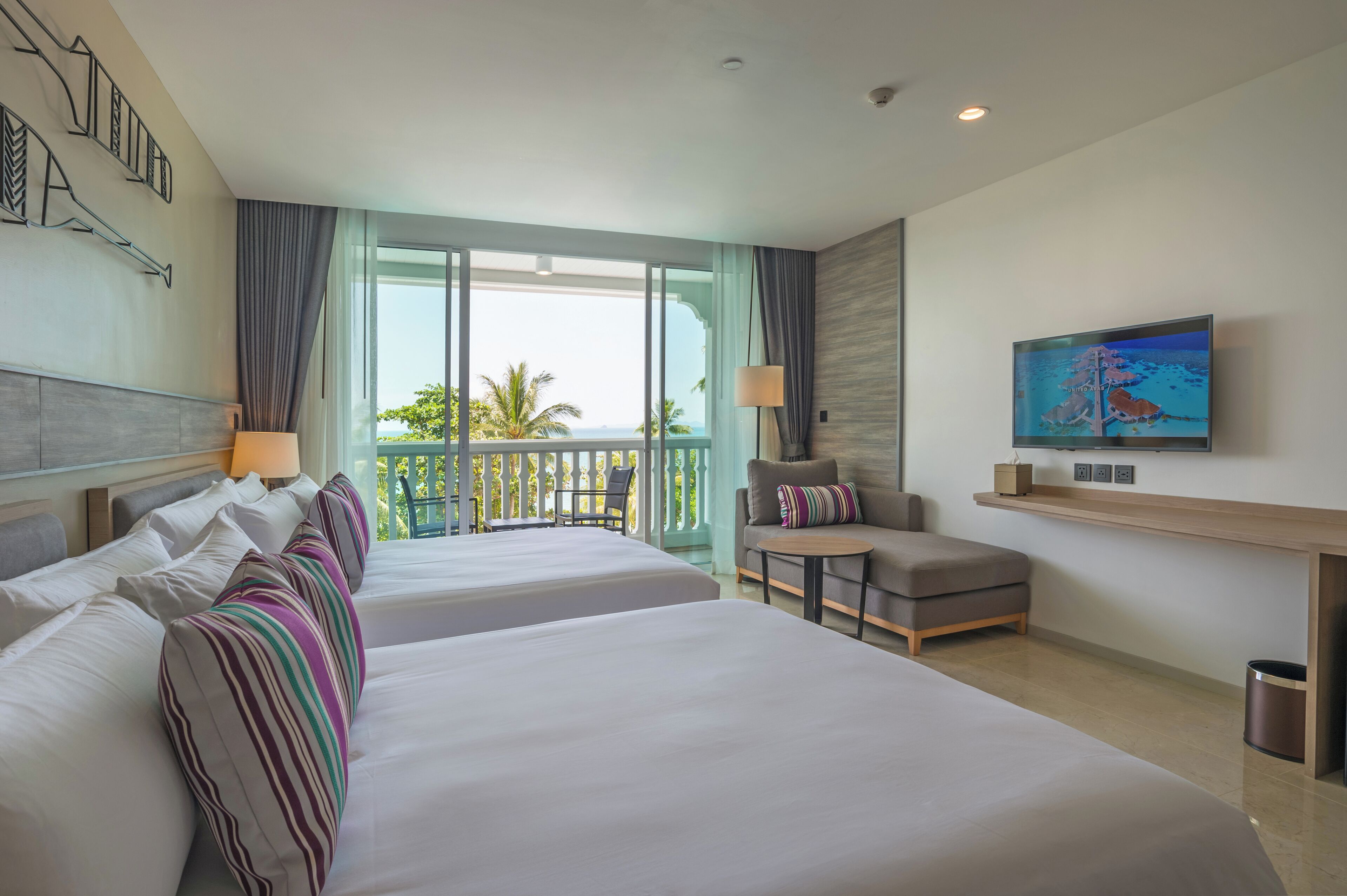 A hotel room with two beds, a sofa, a TV, and a balcony with a view of palm trees.