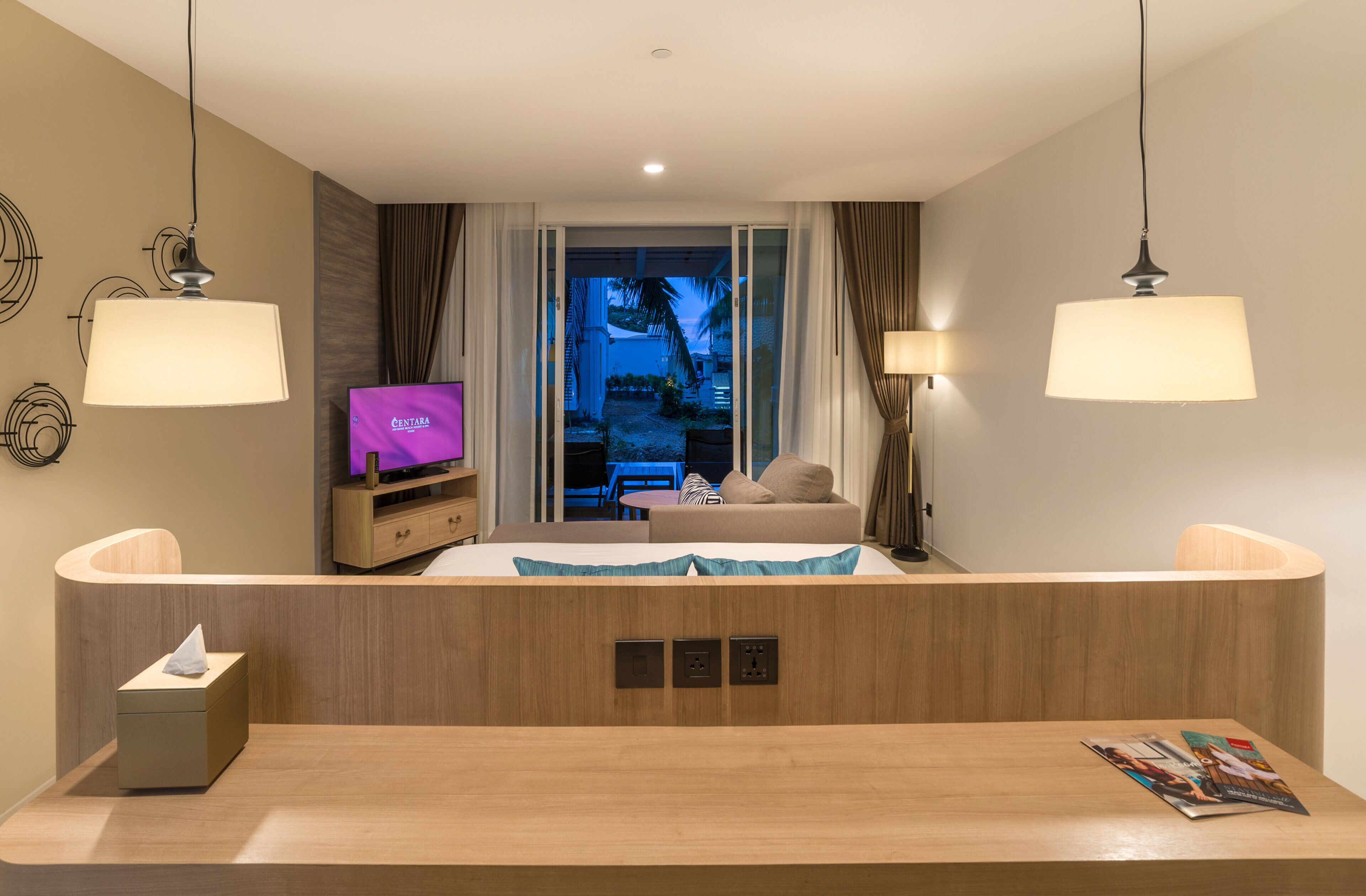 A hotel room with a wooden reception desk, a television, a sofa, and a view of the outdoor area.