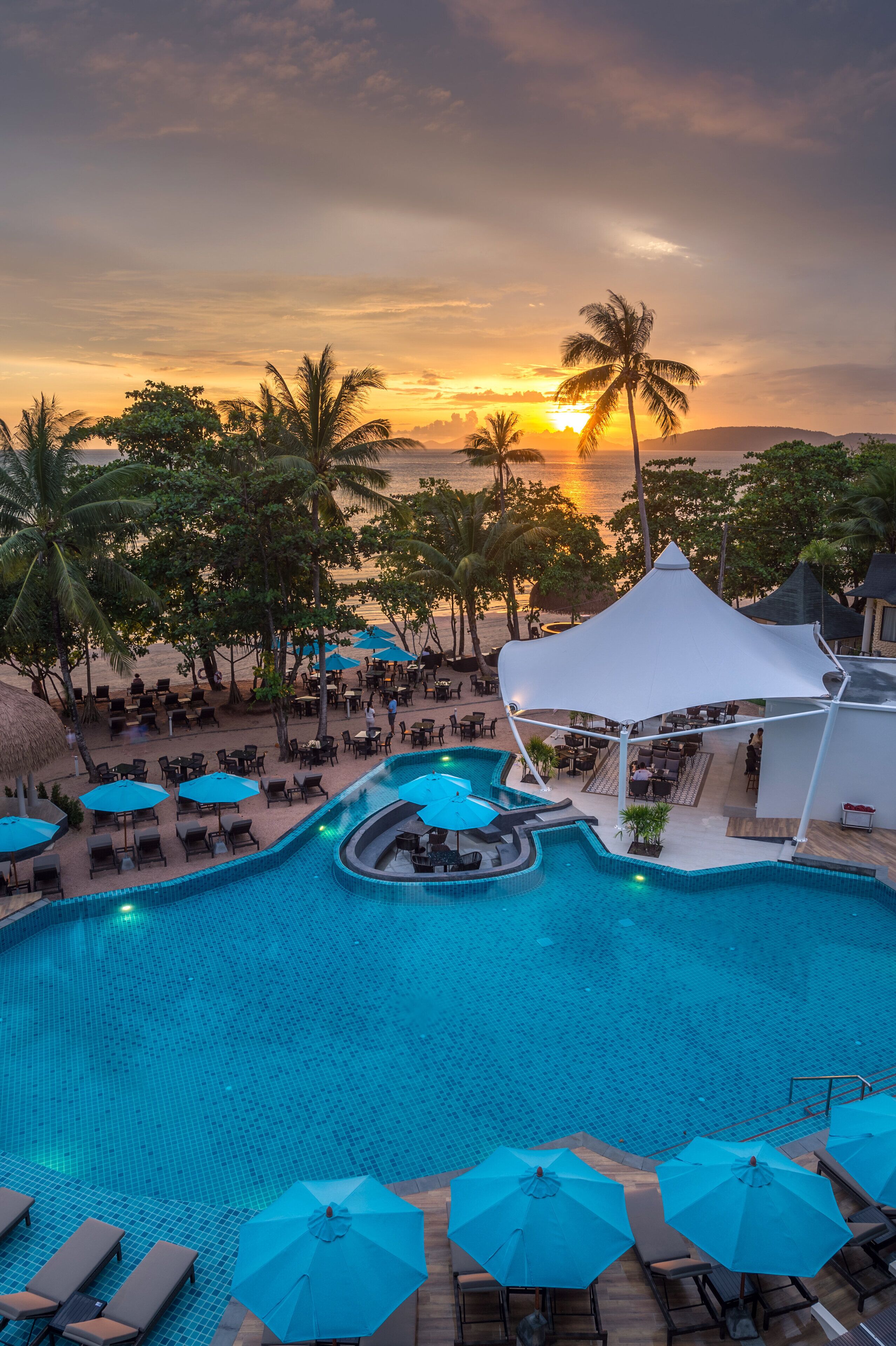 A resort pool area with lounge chairs and umbrellas, surrounded by palm trees and a sunset view.