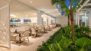 Un comedor interior moderno con sillas y mesas de mimbre, rodeado de exuberante vegetación y un muro de cristal que ofrece vistas del interior.