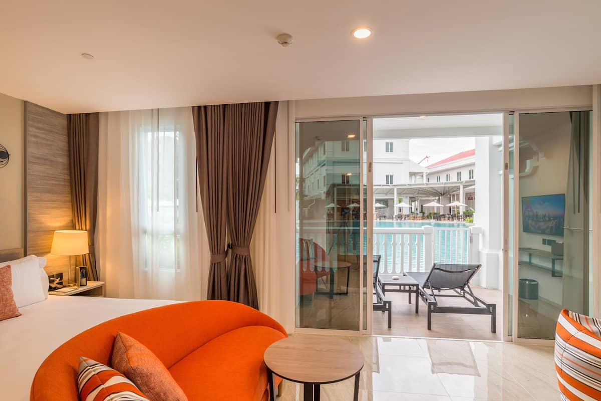 Ein Hotelzimmer mit Bett, einem orangefarbenen Sofa, einem kleinen Tisch und einem Balkon mit Pool und Sitzgelegenheit.