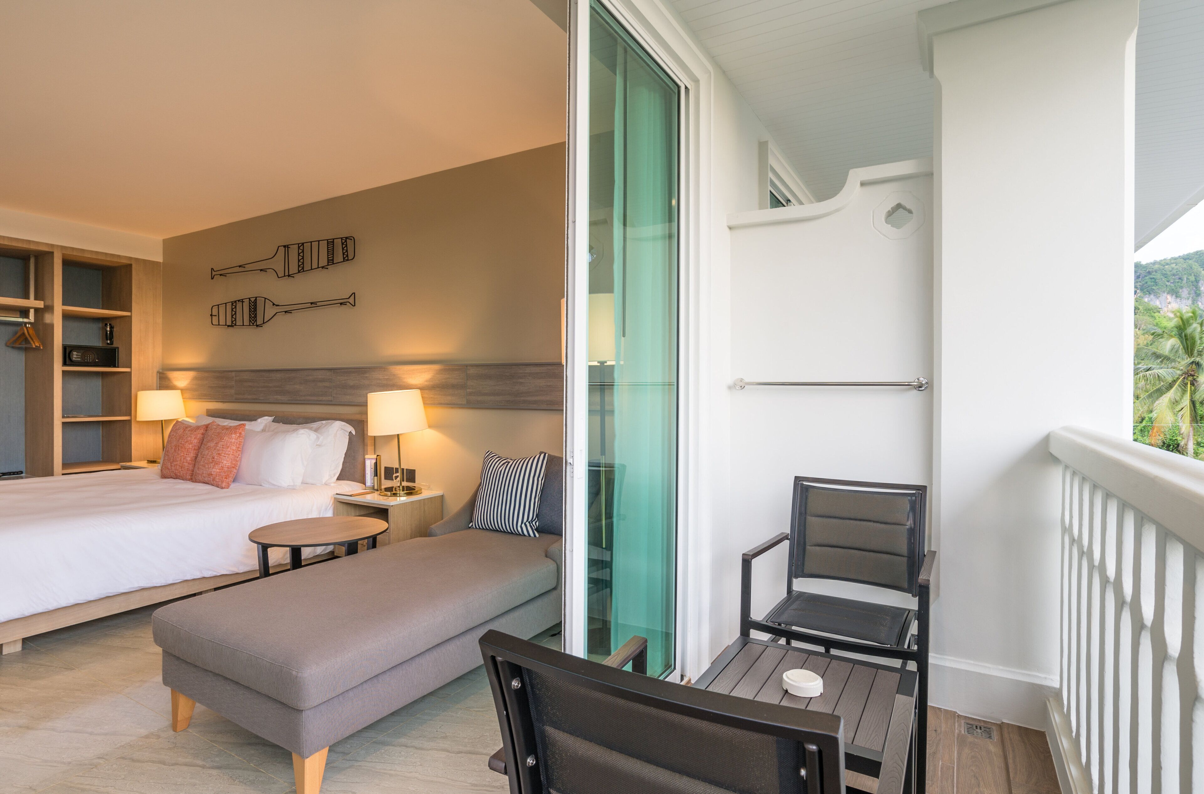 Une chambre d’hôtel avec un lit, un canapé, une petite table et une chaise. Une porte vitrée donne sur un balcon agrémenté d’une balustrade.