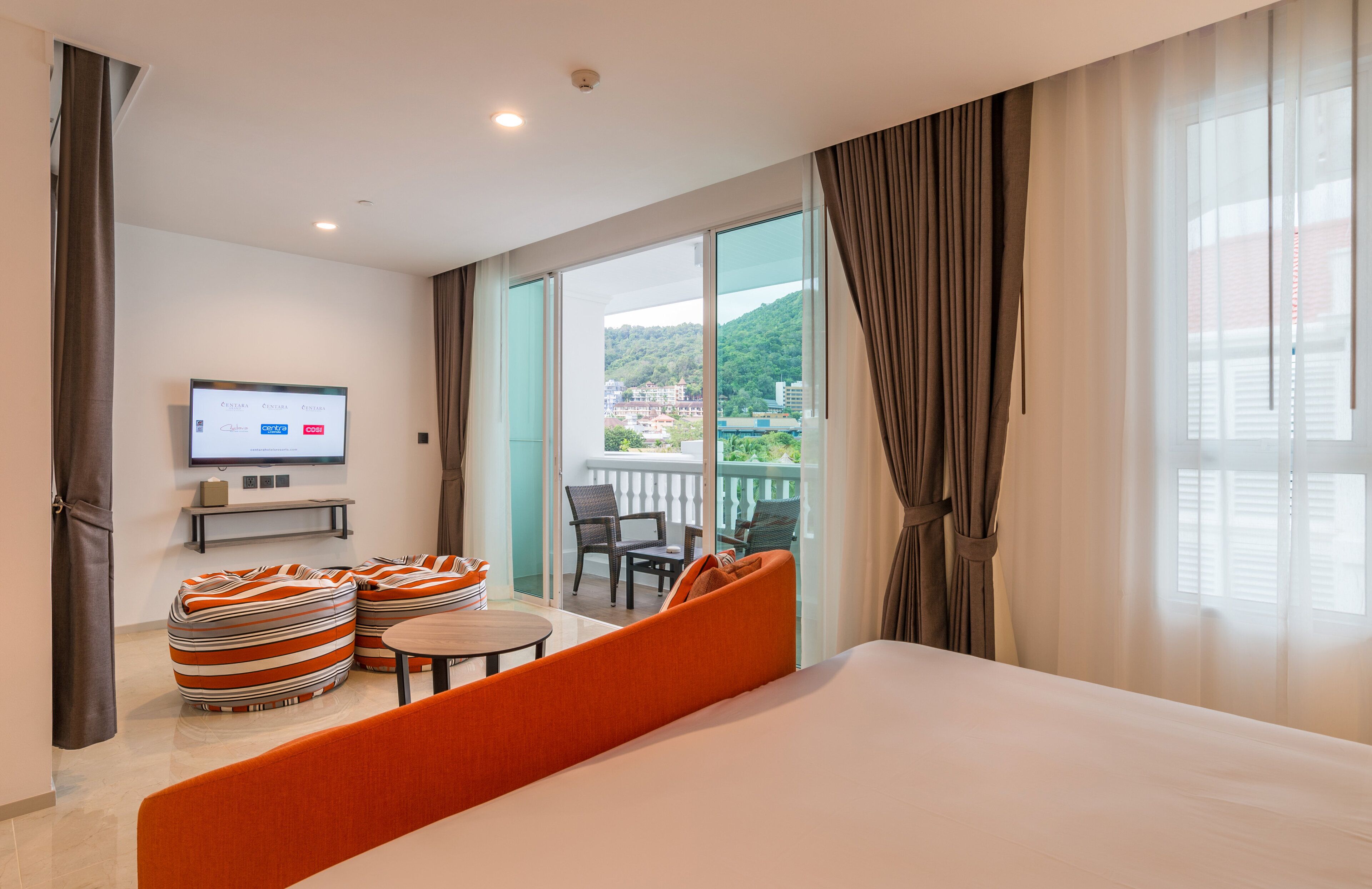 A hotel room with a large bed, a TV on the wall, a balcony with a view of the city, and striped bean bag chairs.