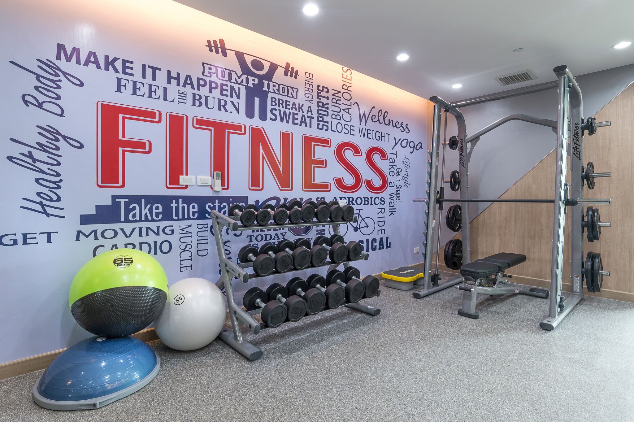 A gym with a wall decorated with fitness-related words and phrases, a rack of dumbbells, a medicine ball, and a bench.