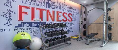A gym with a wall decorated with fitness-related words and phrases, a rack of dumbbells, a medicine ball, and a bench.