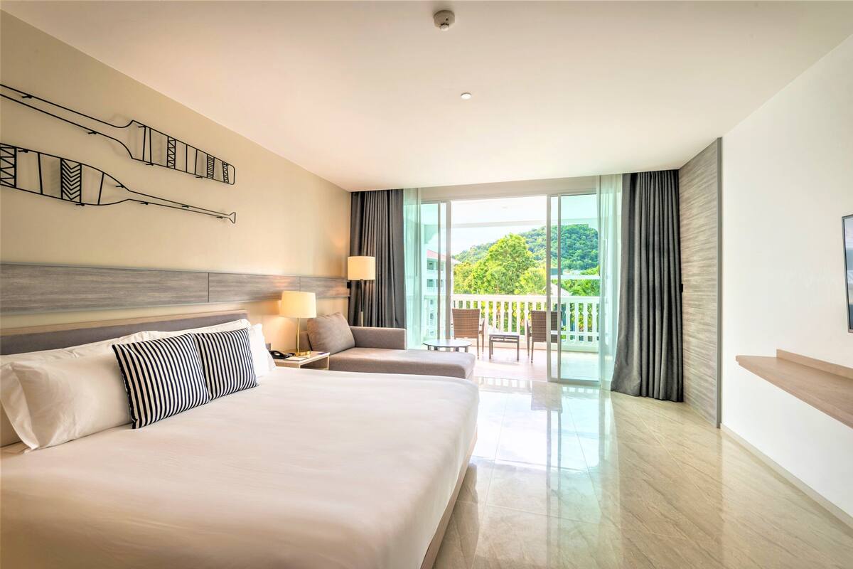 Ein Hotelzimmer mit einem großen Bett, einem Schreibtisch, einem Stuhl und einem Balkon mit Blick auf Grünfläche.