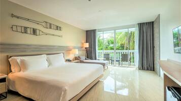 A hotel room with a large bed, a desk, a chair, a balcony with a view of greenery, and a wall-mounted artwork.