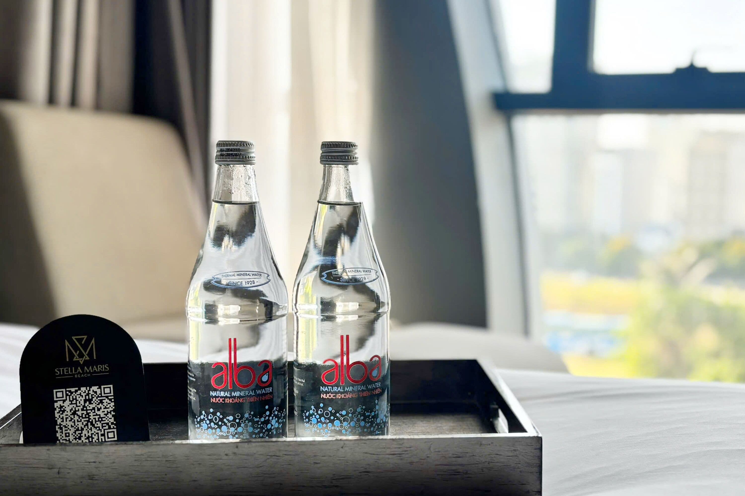 Two bottles of water labeled 'alba' on a tray next to a QR code and a sign with 'STELLA MARIS' branding.