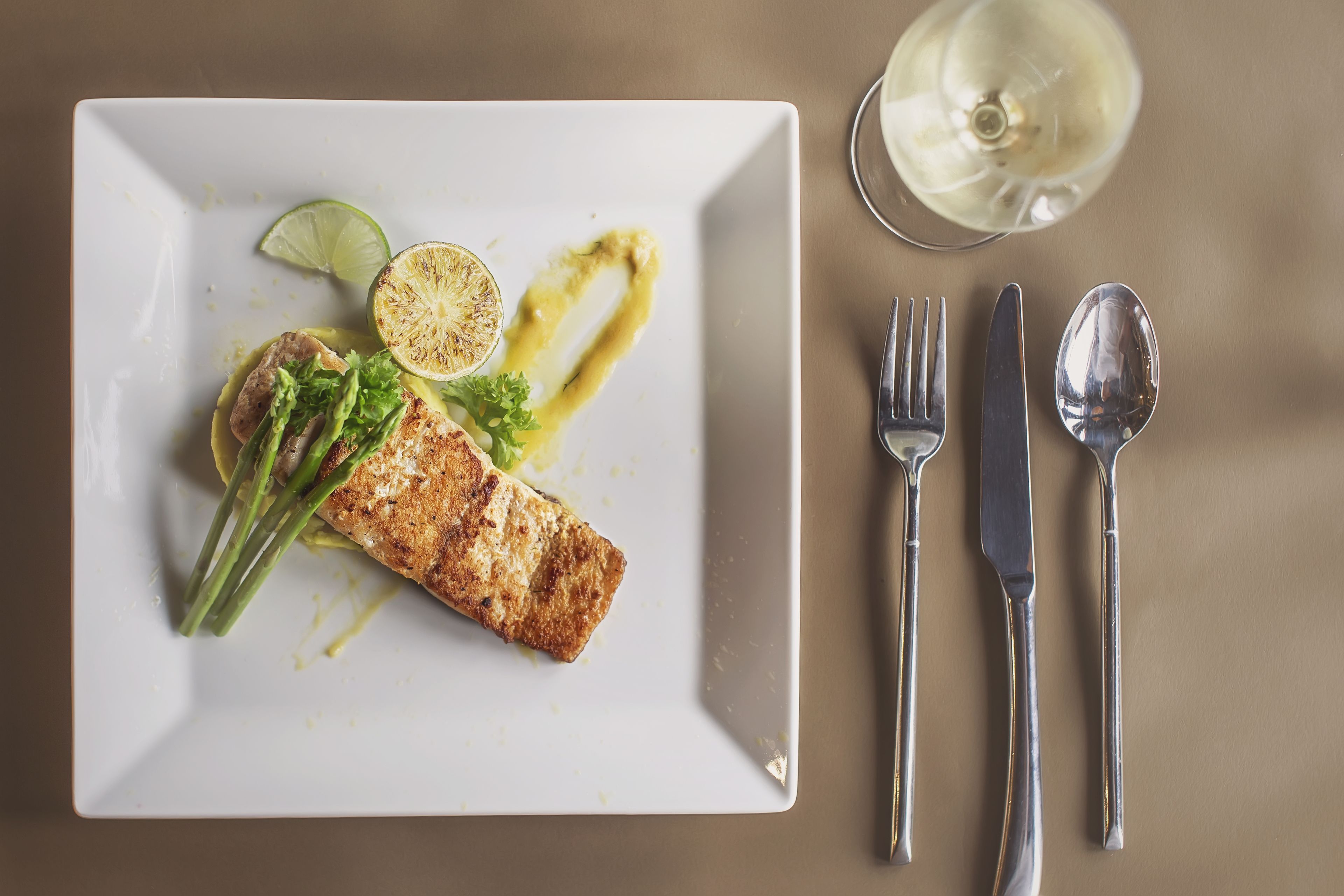 Ein weißer Teller mit gegrilltem Fisch, grünem Gemüse und einer Limettenscheibe auf einem beigefarbenen Tisch.