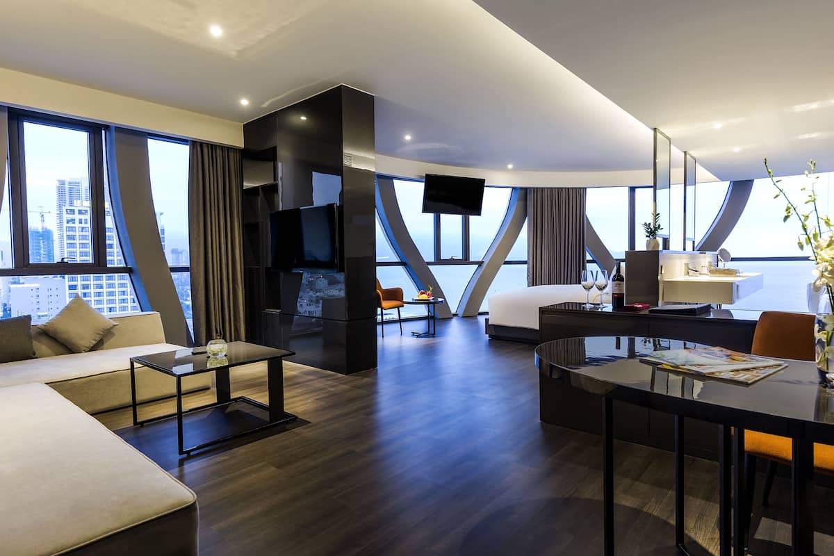 A modern hotel room with a large window, a sofa, a dining table, and a TV.