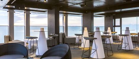 A modern lounge area with seating arrangements, large windows, and a view of the sea.
