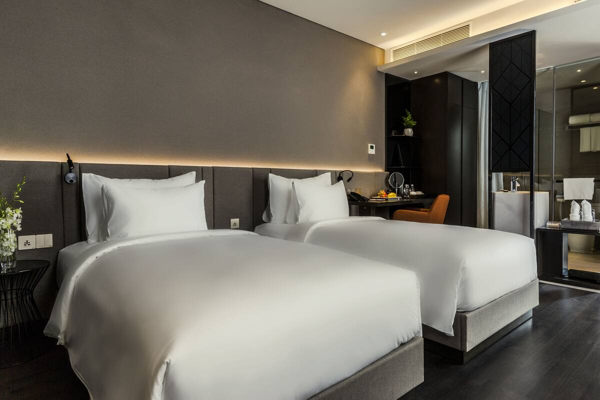 A modern hotel room with two large beds, a desk, a chair, and a bathroom visible through a glass partition.