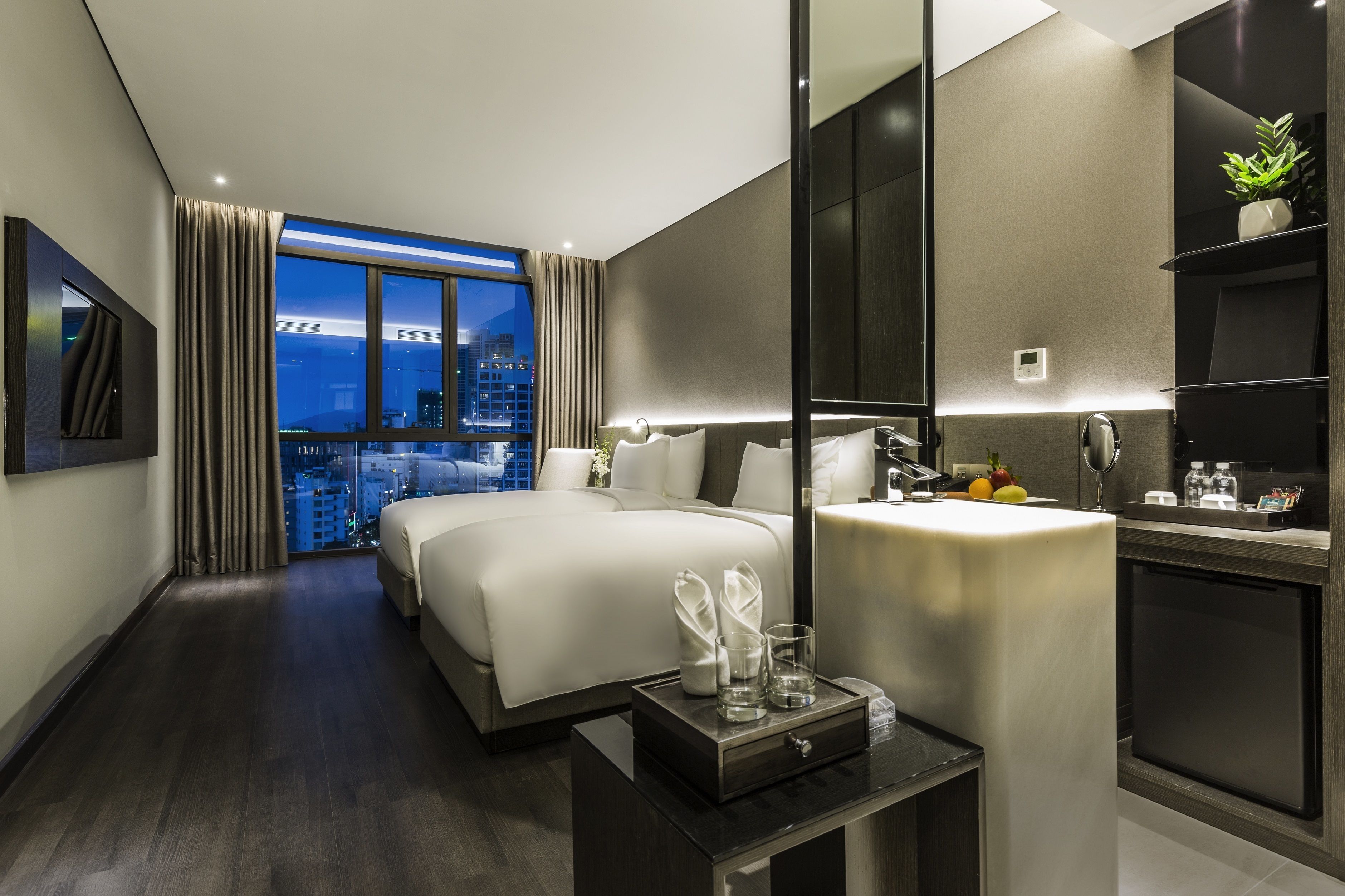 Ein modernes Hotelzimmer mit einem großen Bett, einem Nachttisch mit Lampe, einem kleinen Kühlschrank und Blick auf die beleuchtete Stadt bei Nacht.