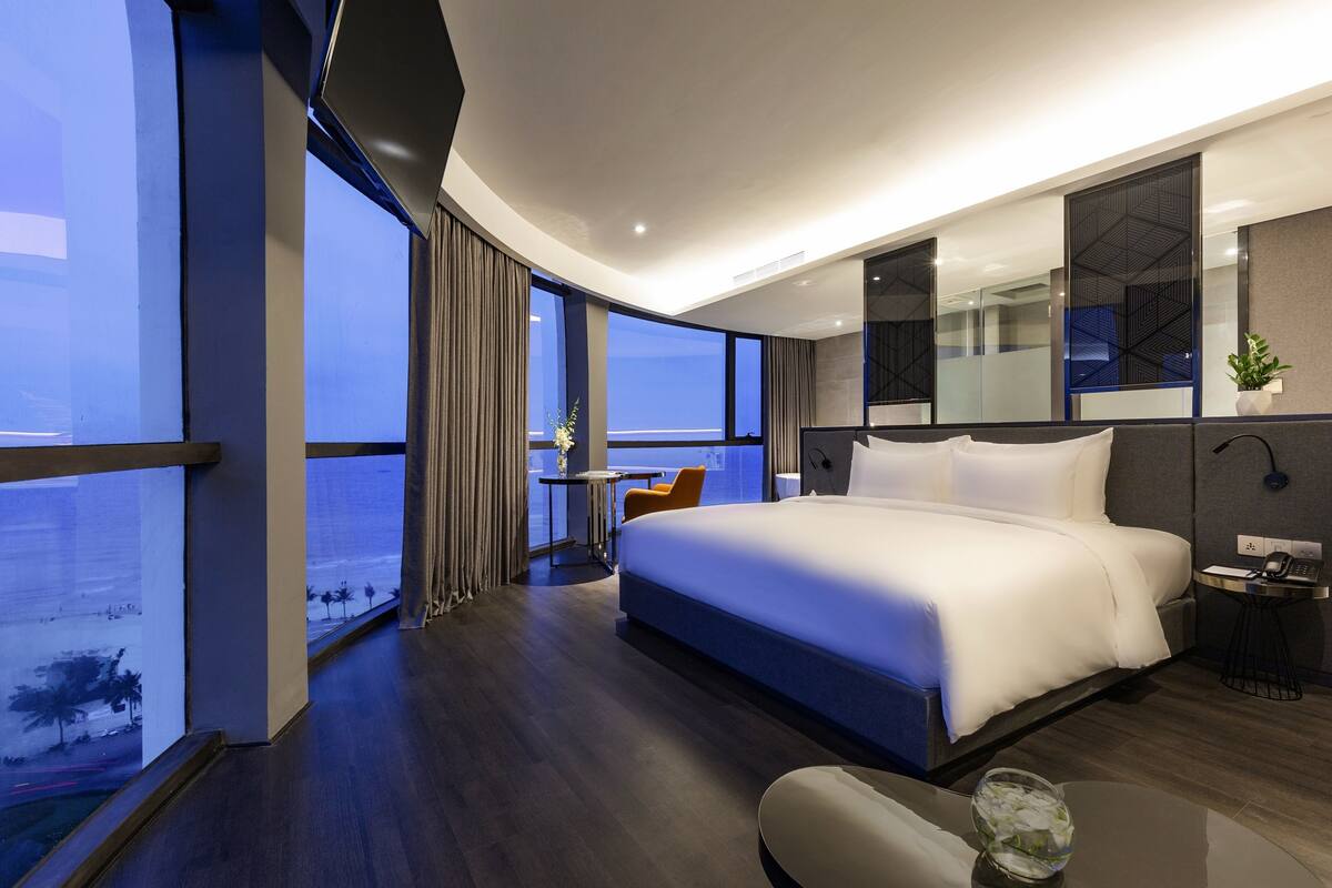 A modern hotel room with a large bed, a small table, a chair, and a large window offering a view of the city.