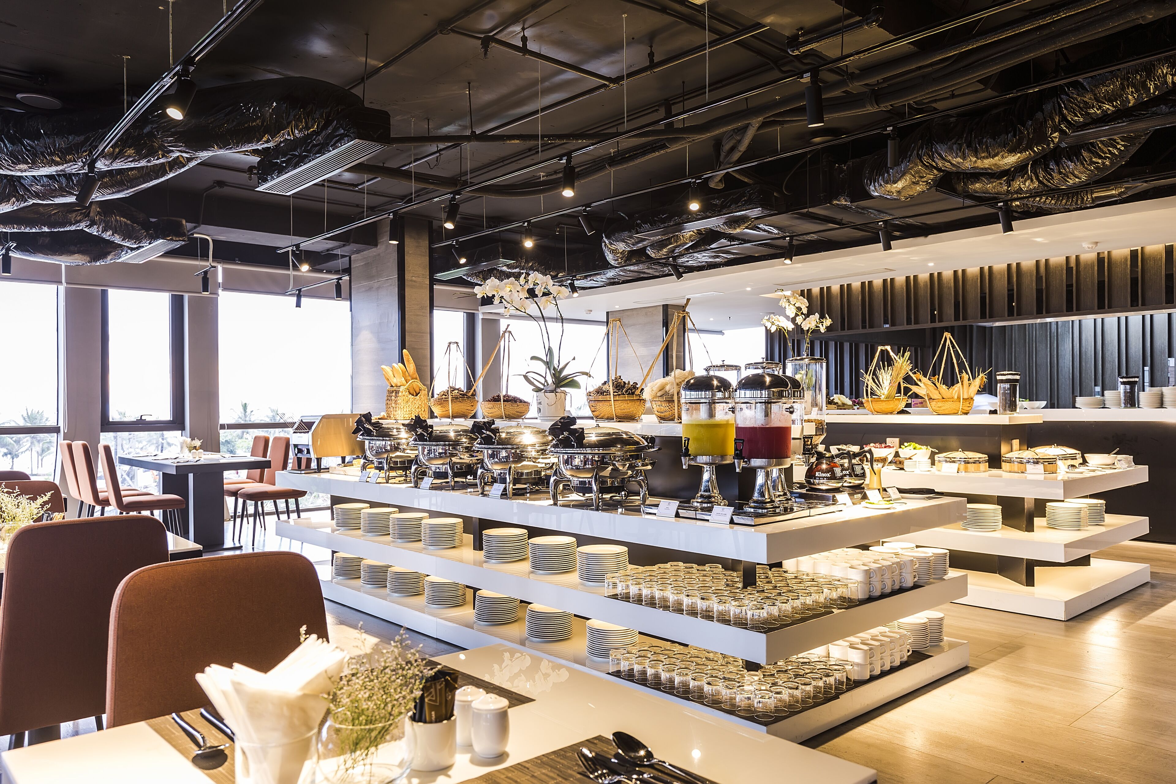 A modern restaurant with multiple dining tables and chairs, a buffet area with various food items, and a view of the city through large windows.