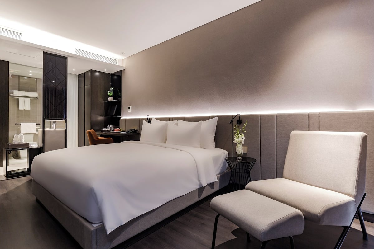A modern hotel room with a large bed, a bedside table, a chair, and a small desk with a lamp.