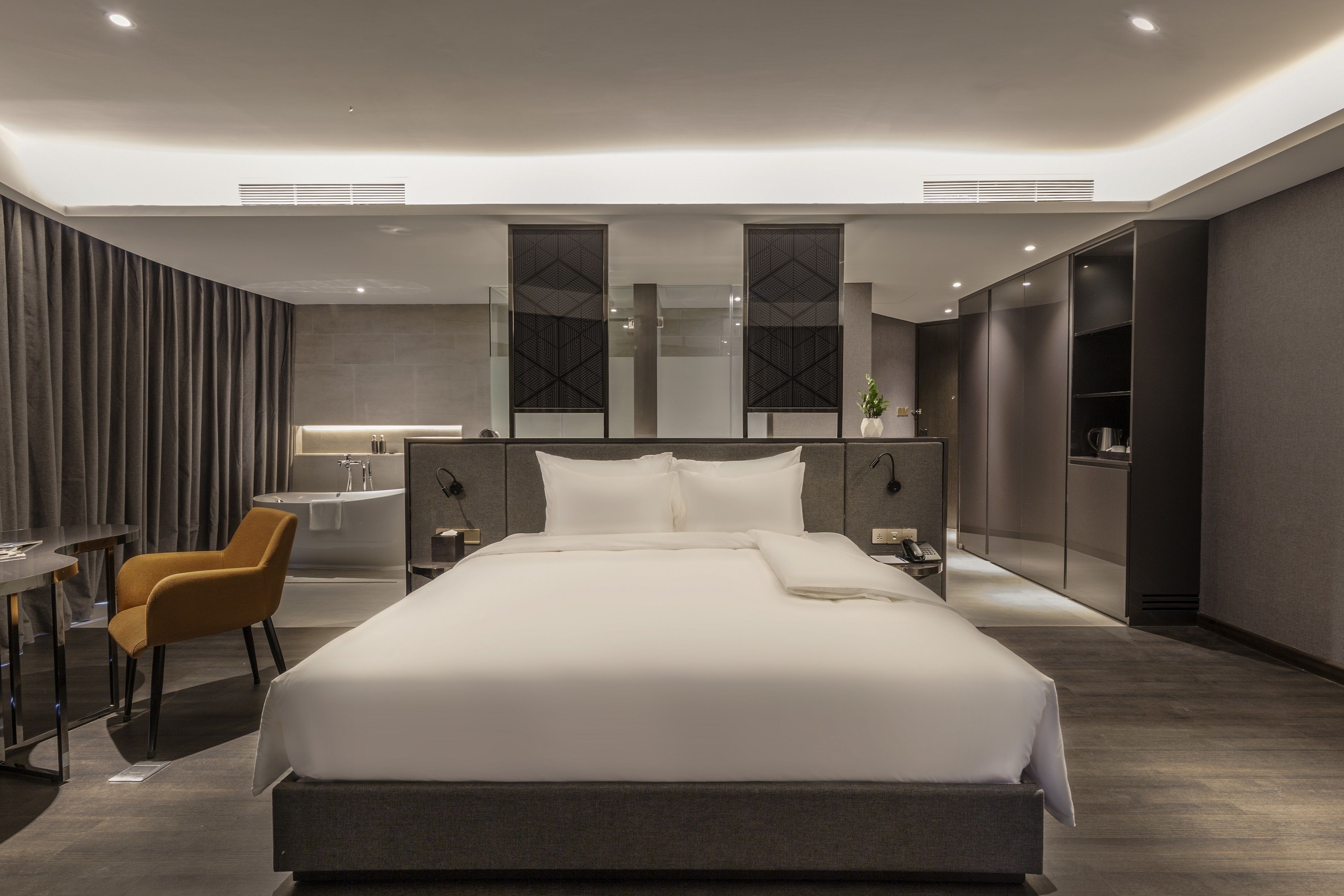 A modern hotel room with a large bed, a desk, a chair, a vanity with a mirror, and a small plant.
