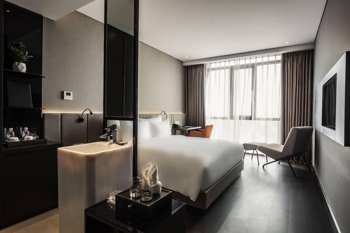 A modern hotel room with a large bed, a desk, a chair, a vanity with a mirror, and a potted plant.