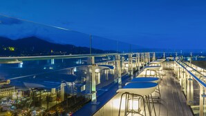 Ein moderner Außenbereich zum Essen mit Glasgeländern und Metallstühlen, mit Blick auf eine Stadtlandschaft, inklusive Brücke und Bergen im Hintergrund.