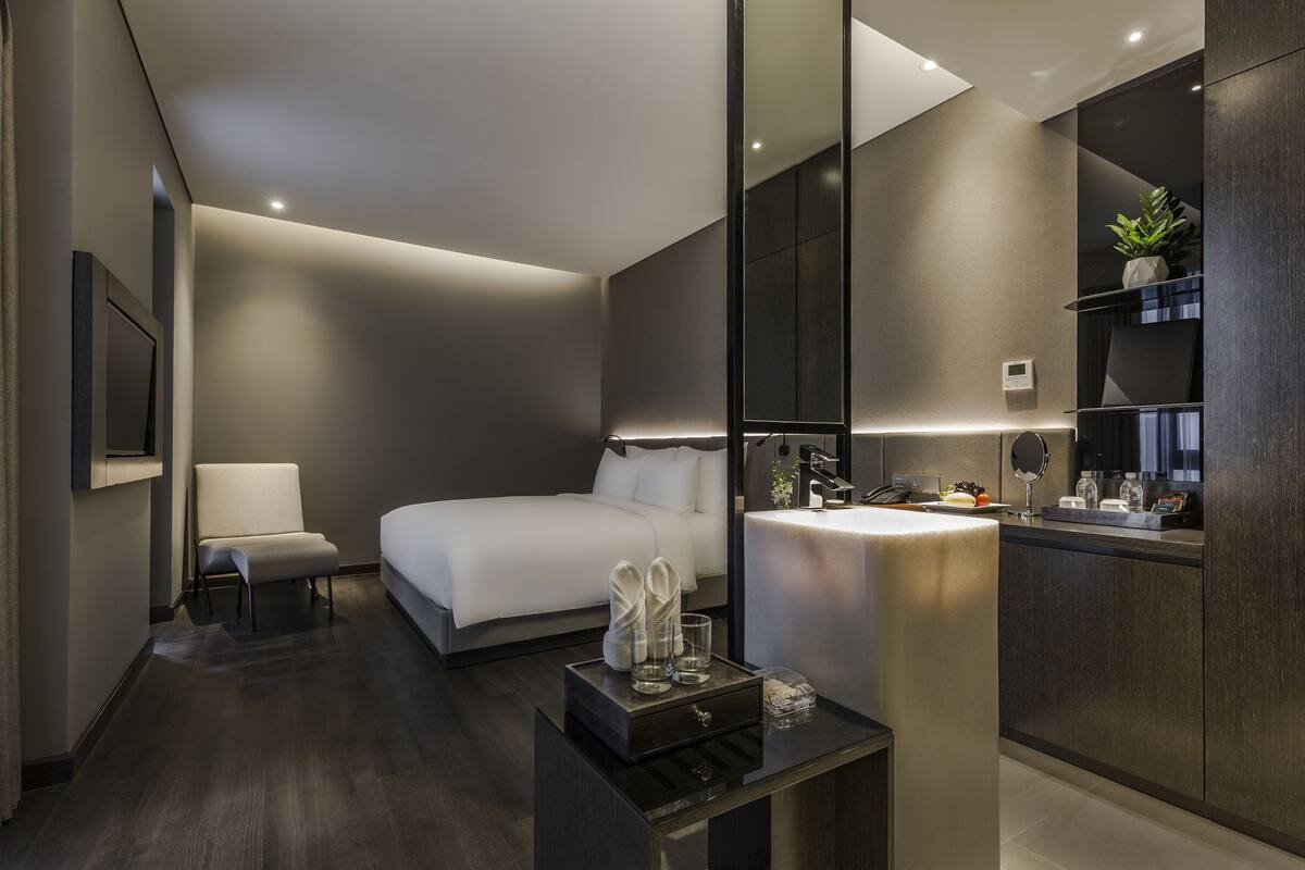 A modern hotel room with a bed, a bedside table with a mirror, a chair, and a small table with toiletries.