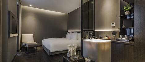 A modern hotel room with a bed, a bedside table with a mirror, a chair, and a small table with toiletries.