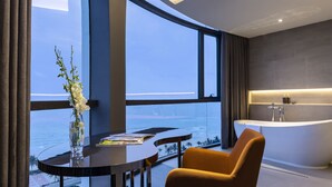 Ein modernes Badezimmer mit einer freistehenden Badewanne, einem großen Fenster mit Meerblick und einem Tisch mit Blumen in einer Vase.