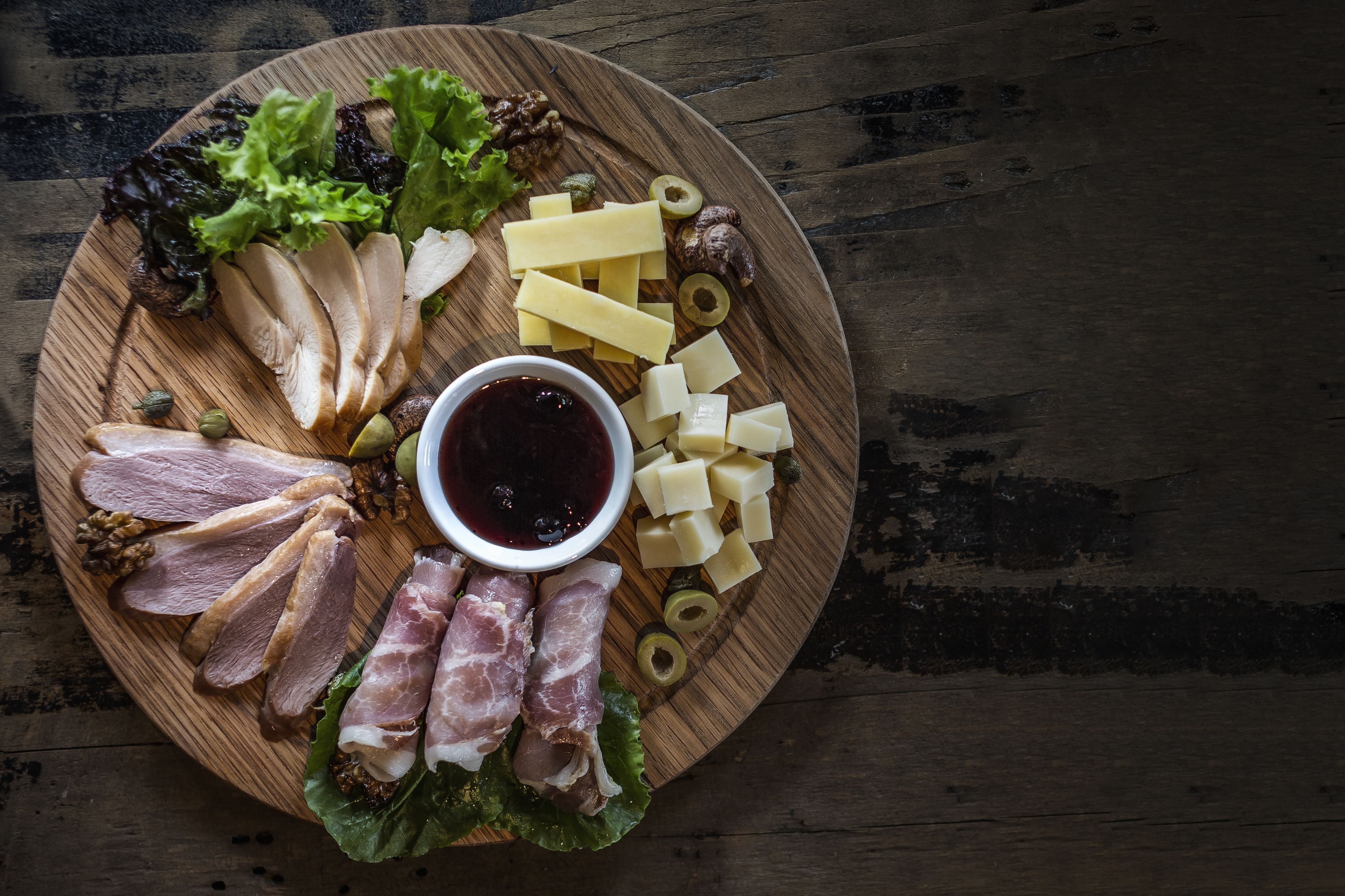 Una tabla de madera con carnes cortadas, queso, aceitunas y un tazón de salsa.