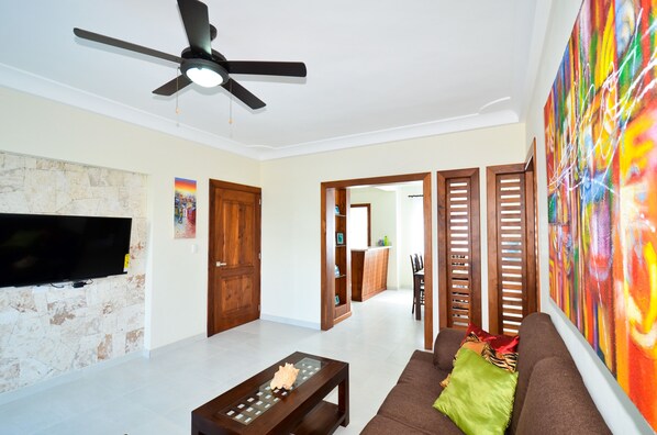 A spacious living room with a flat-screen TV, a ceiling fan, a stone wall, wooden doors, and a colorful abstract painting. -  2 Bedroom, 2 Bath Ground Level Condo (Punta Cana)