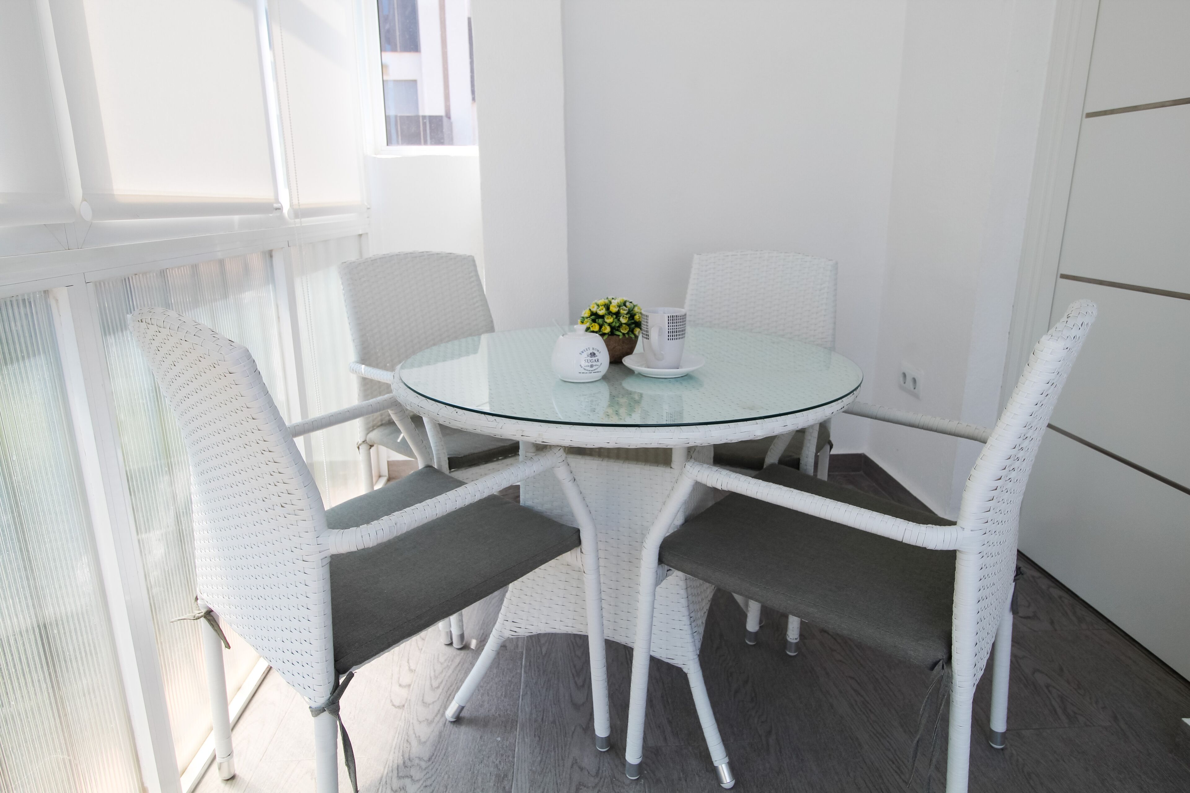 A round glass table with white wicker chairs, a small potted plant, and a white mug on a white tablecloth.