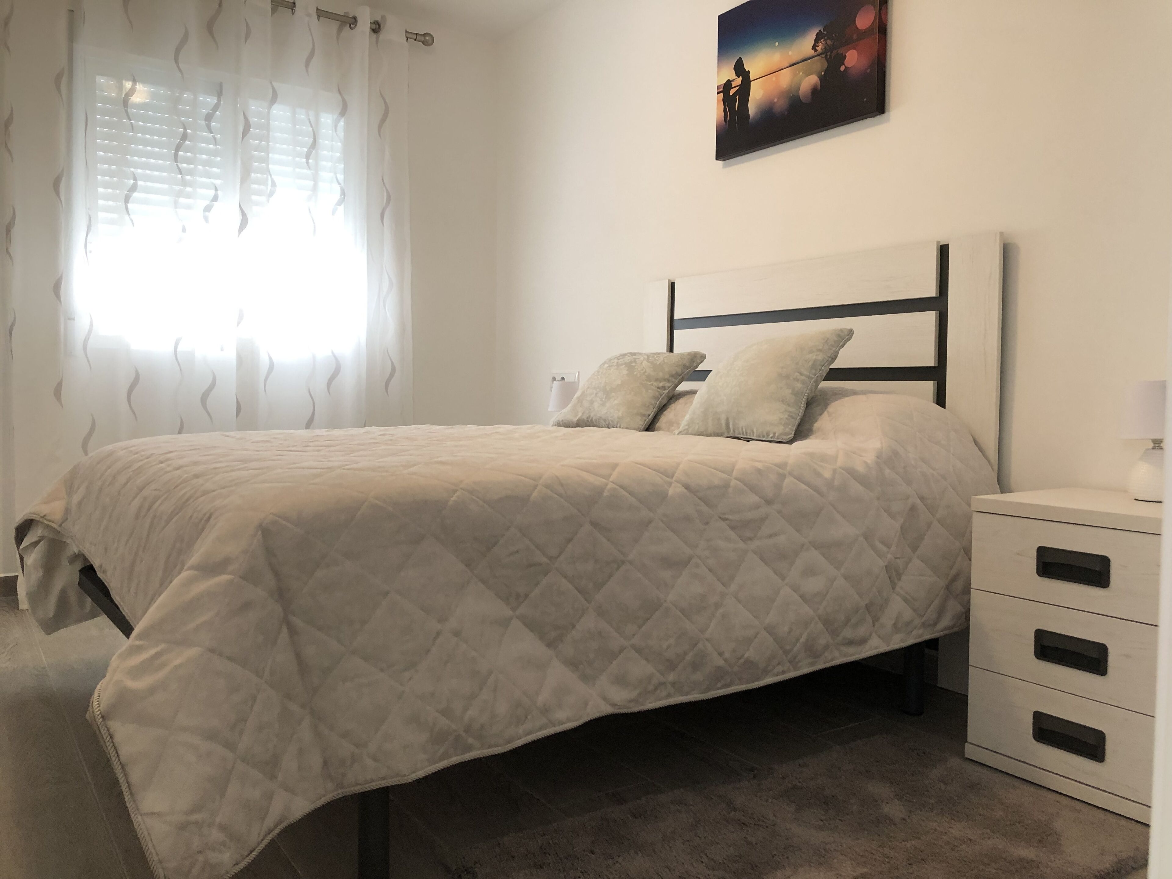 A neatly made bed with a quilted cover, a bedside table with a lamp, and a painting on the wall.
