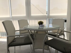 Ett runt glasbord omgivet av vita flätade stolar med mörka kuddar. En liten krukväxt med gula blommor står på bordet.