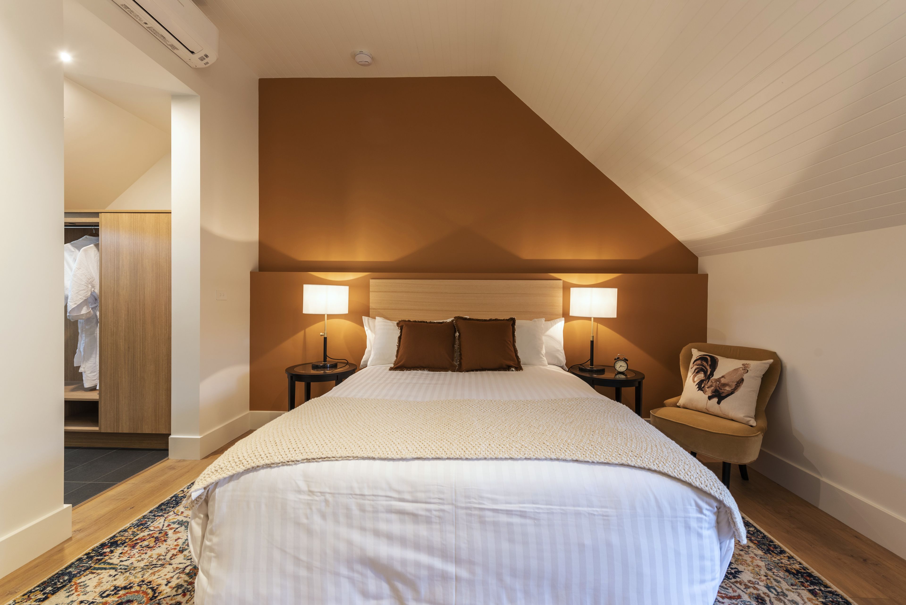 A bedroom with a large bed, two bedside lamps, a nightstand, a chair with a cushion, and a wooden wardrobe.