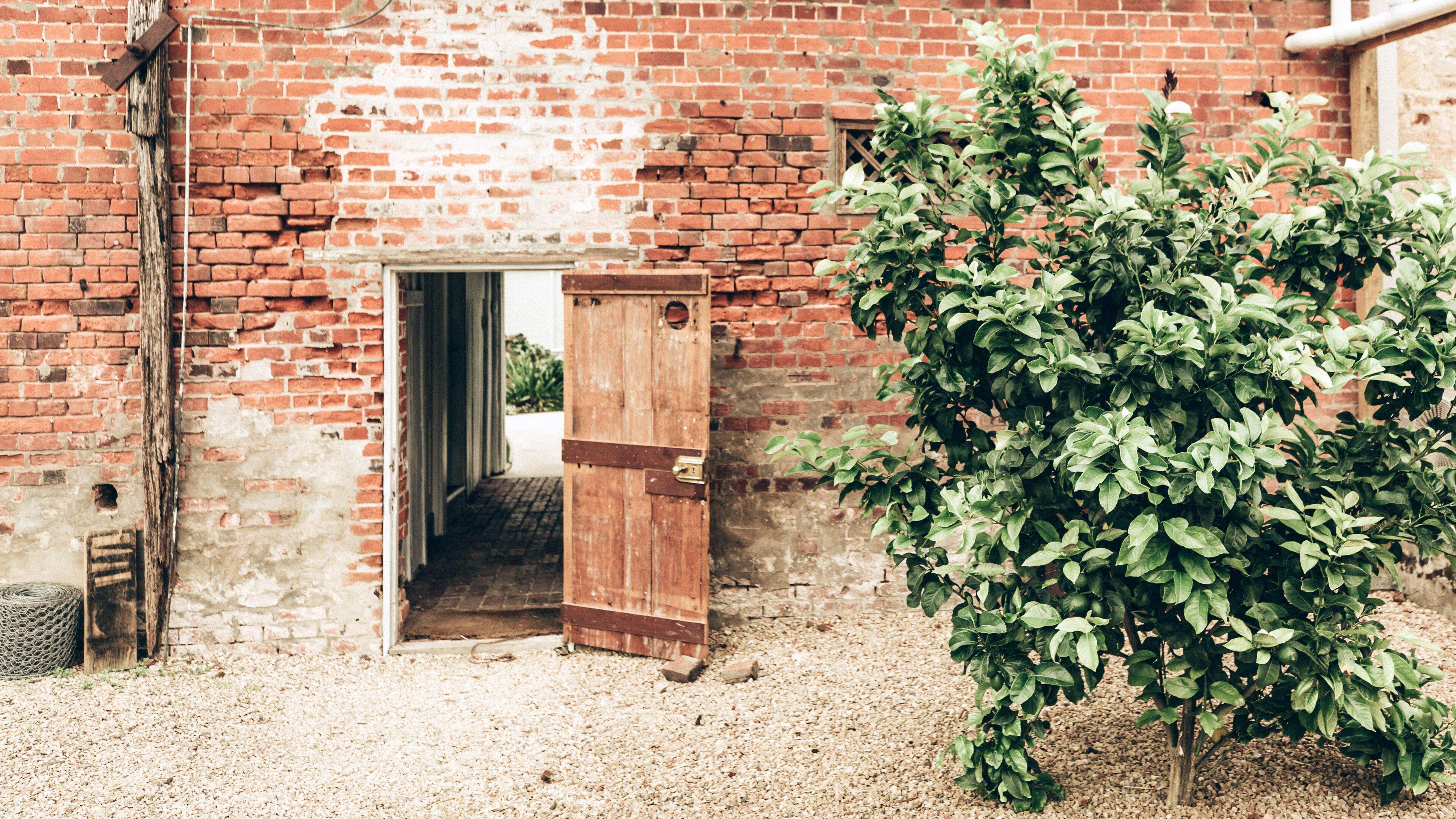 An open wooden door leading to a brick building, surrounded by gravel and a green bush.